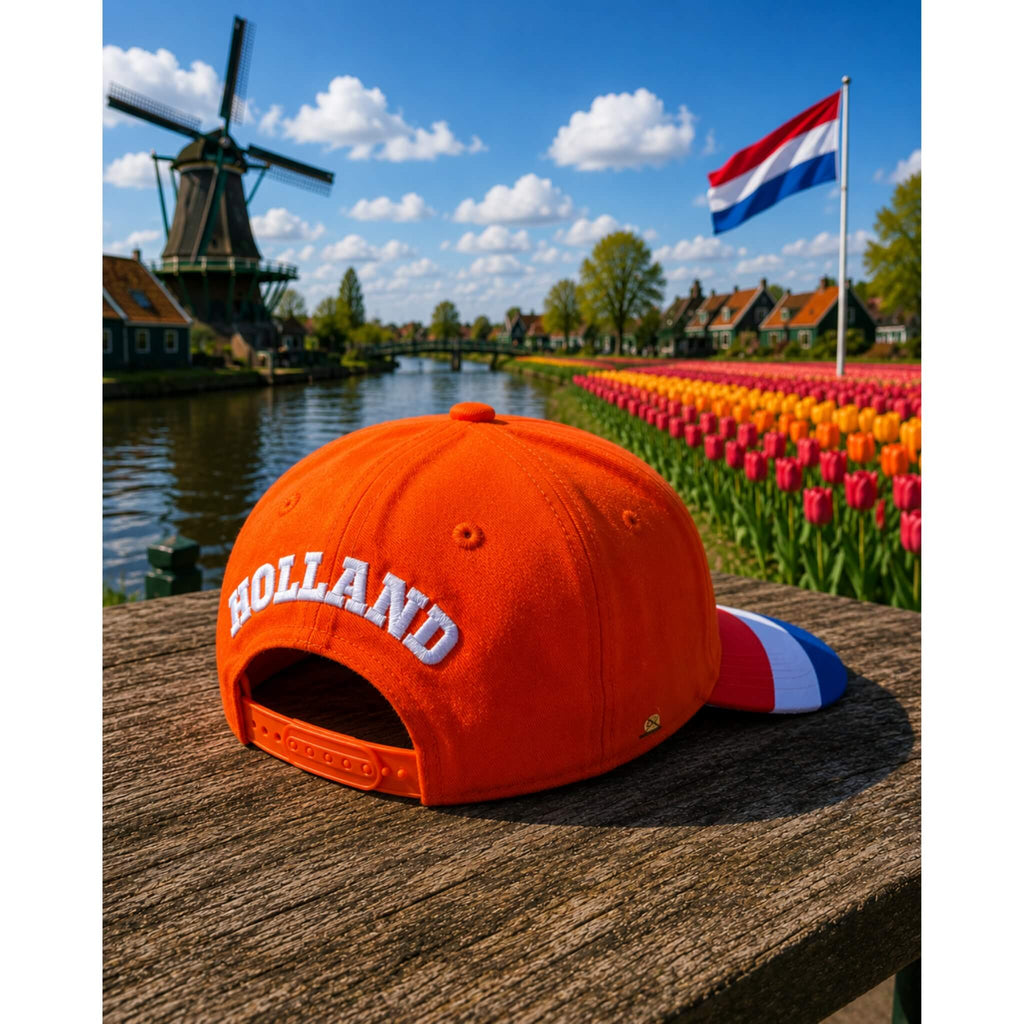 Oranje Holland supporterspet van BNationX met geborduurd “HOLLAND” logo en rood-wit-blauwe details, afgebeeld bij typisch Nederlands landschap met molen, tulpenveld en Nederlandse vlag.