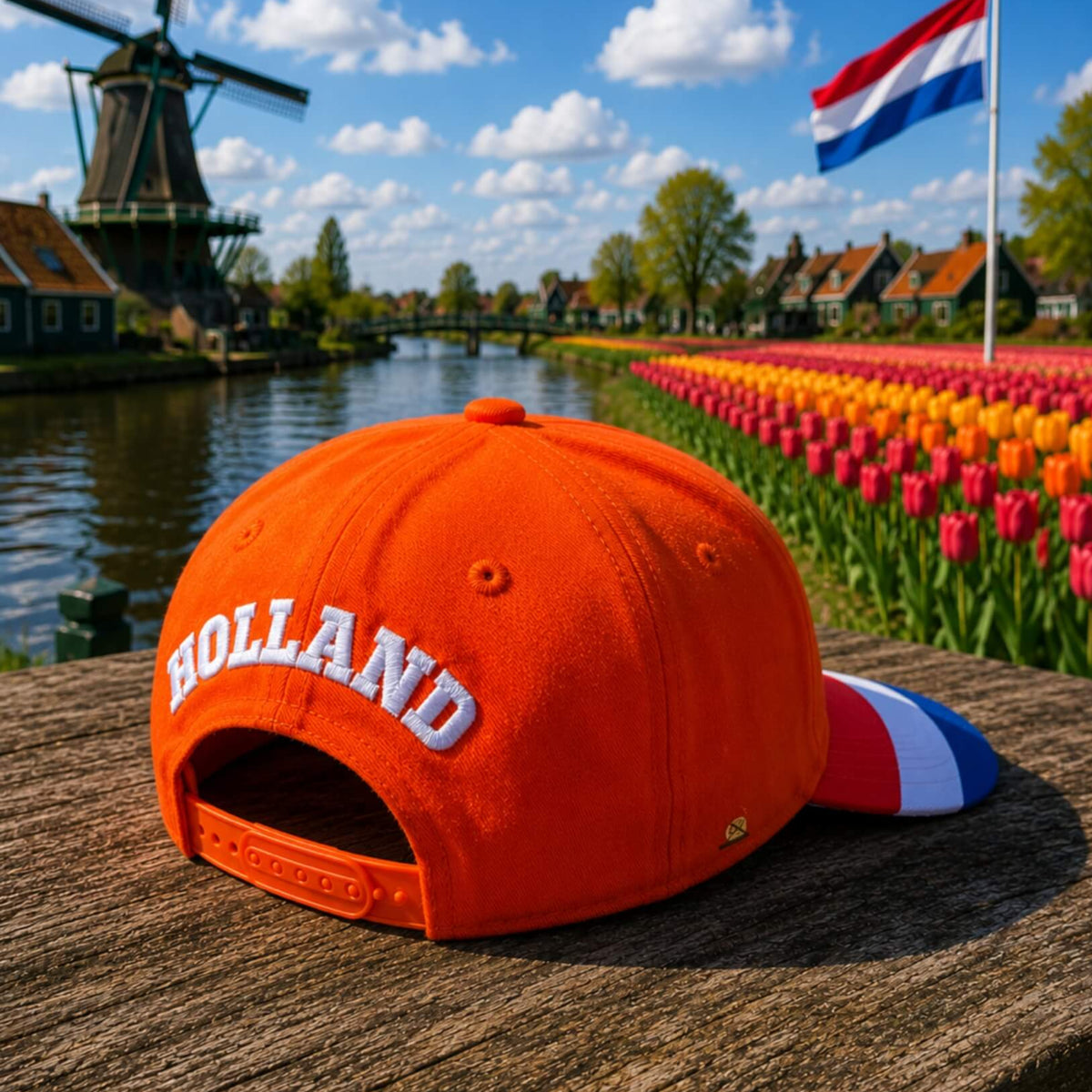 Oranje Holland supporterspet van BNationX met geborduurd “HOLLAND” logo en rood-wit-blauwe details, afgebeeld bij typisch Nederlands landschap met molen, tulpenveld en Nederlandse vlag.
