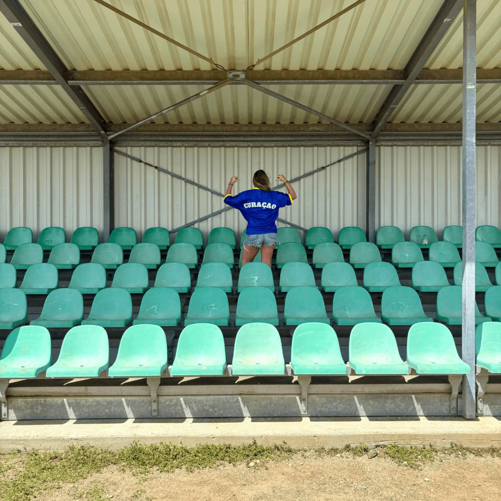 Supporter draagt een blauw Curaçao supportersshirt van BNationX in een voetbalstadion en toont trots de achterkant met “CURAÇAO” op de rug. Uniseks supporterskleding geïnspireerd op de Curaçaose vlag voor WK voetbal fans.